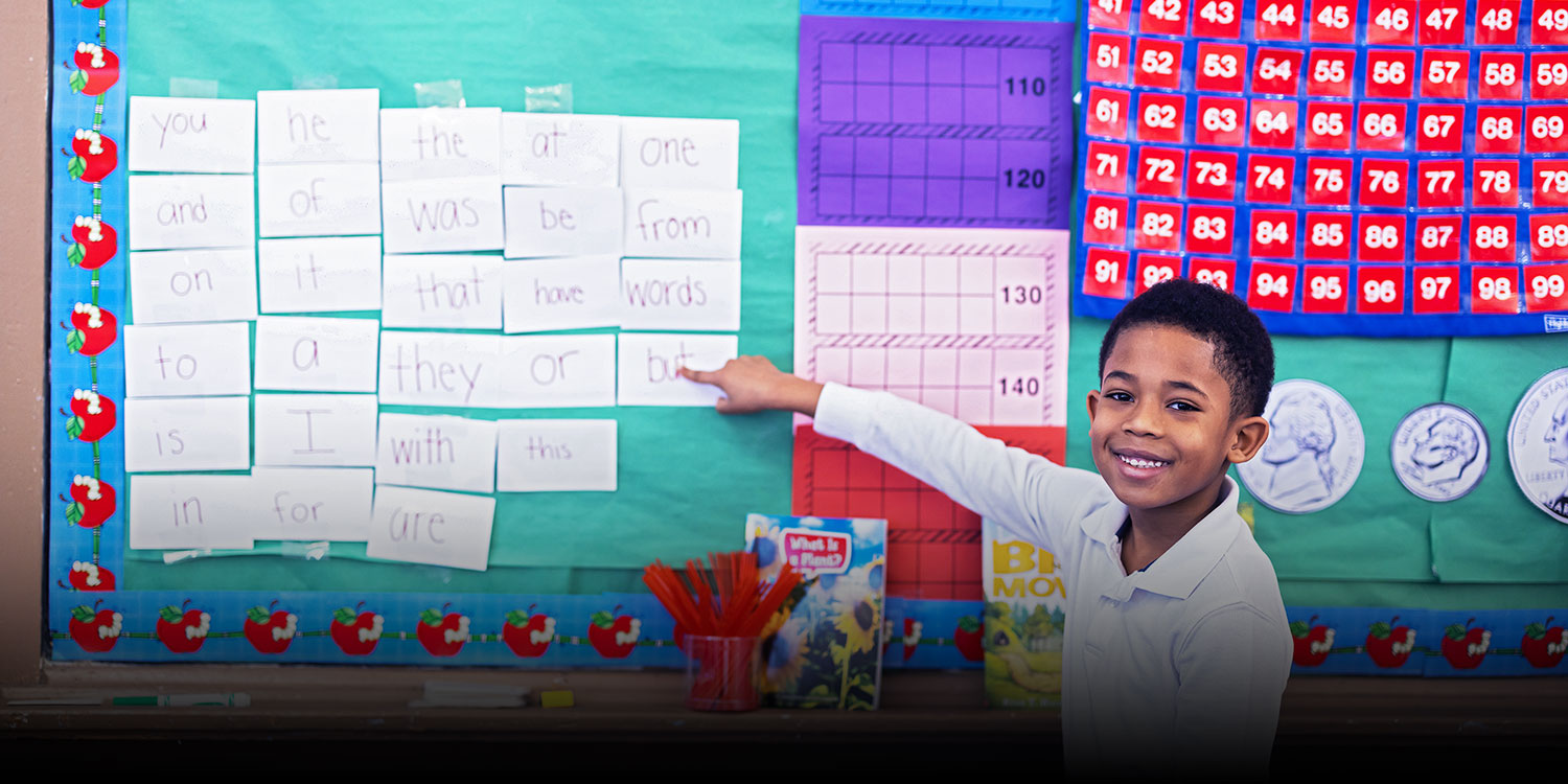 Student smiling and pointing to sight words displayed in a classroom.